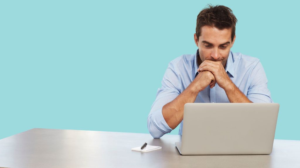 Man sitting at a desk looking thoughtfully at a laptop, focused on work or decision-making.